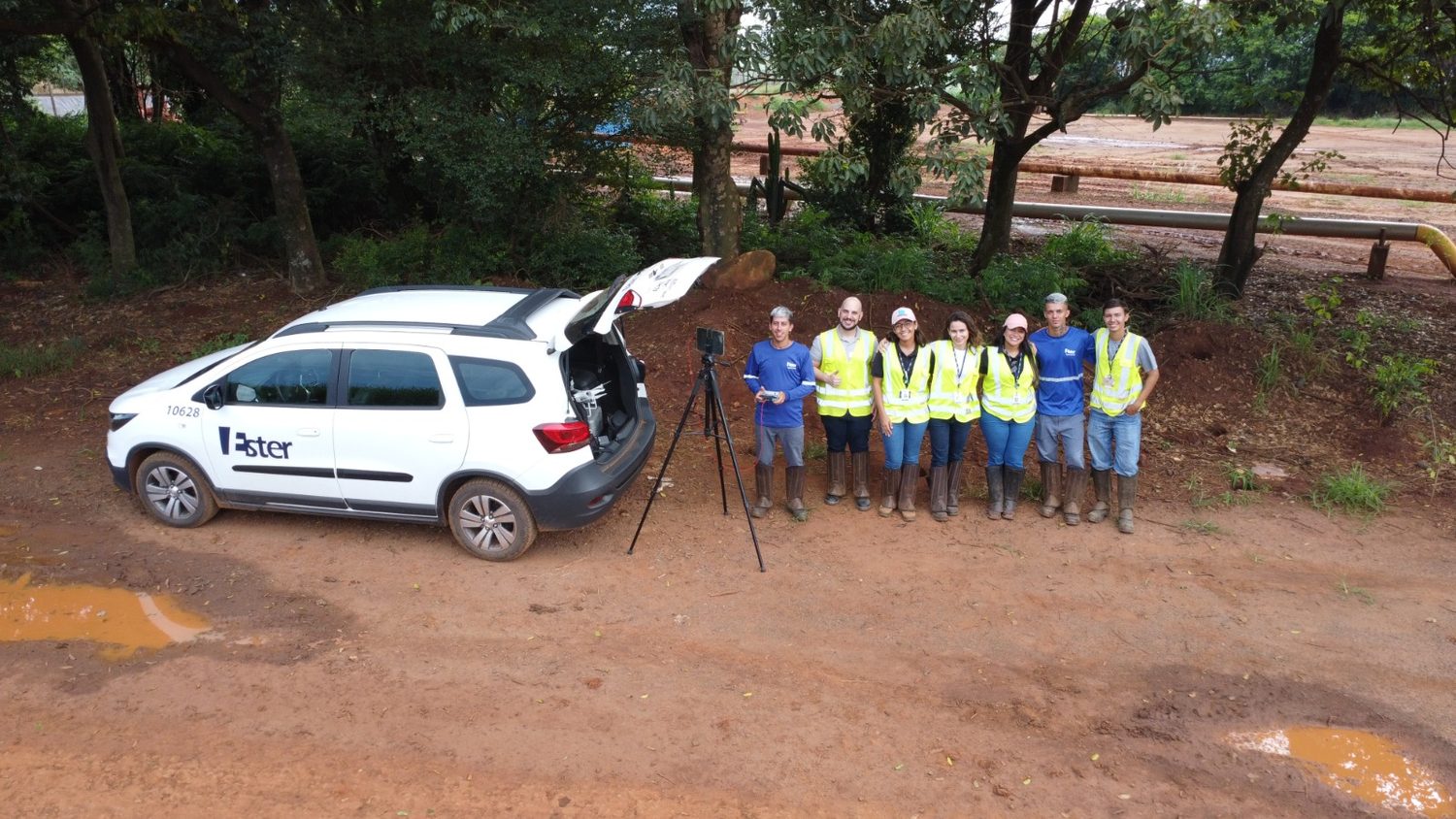 Imagem aérea de equipe de campo com colete refletivo durante capacitação com drone, com tubulação industrial ao fundo